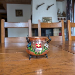 Op de afbeelding: Een kleine, koperkleurige pot met zwarte handvatten en poten. De pot is versierd met kleurrijke bloemmotieven en een rood-wit Zwitsers kruis embleem. De pot staat op een houten tafel.