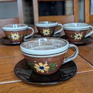 Op de afbeelding: Set van vier keramische kopjes en schotels. De kopjes zijn bruin met een witte rand en een bloemenontwerp in geel, blauw en groen. De schotels zijn donkerbruin. De set wordt gepresenteerd op een houten tafel.