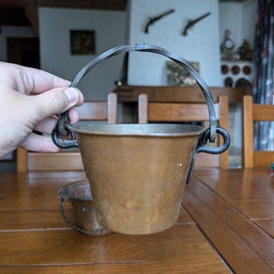 May include: A small, antique copper bucket with a curved metal handle. The bucket has a rounded body and is held by a hand. A second, smaller bucket is visible in the background. The background includes a wooden table and chairs.