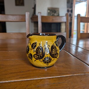 Pode incluir: Um jarro de cerâmica amarela com uma alça e borda marrom escuro. O jarro é decorado com desenhos florais e de borboletas pretas. Está sobre uma mesa de madeira.