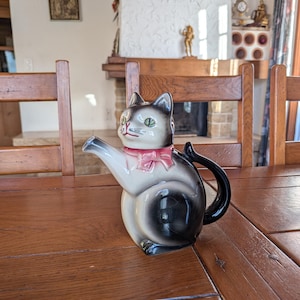 May include: A ceramic cat teapot in black, white, and grey, with a pink bow and a black handle. The teapot has green eyes and a spout. It is placed on a wooden table. The teapot is a decorative item.