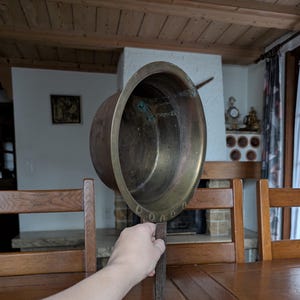 May include: An antique brass pot with a long handle. The pot has a wide, rounded bowl and a dark, aged patina. The handle is made of a darker metal and is attached to the pot with rivets. The pot is held up by a person's hand.