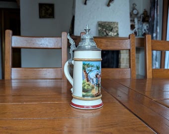 Könnte beinhalten: Ein weißer Bierkrug aus Keramik mit einer detaillierten Illustration eines Mannes auf einem Pferd. Der Krug hat einen Metalldeckel und einen Griff mit roten und goldenen Verzierungen. Der Krug steht auf einem Holztisch.