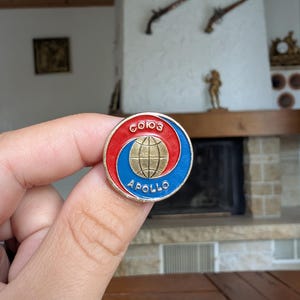 May include: A round pin with a red and blue spiral design, featuring a gold globe in the center. The pin has the words "СОЮЗ" and "APOLLO" in white lettering. The pin is held in a hand.