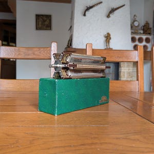 May include: A vintage, silver-toned metal and wood accordion-style musical instrument, resting on a green box. The instrument has intricate detailing and a decorative design. The box is a deep green with a textured surface.