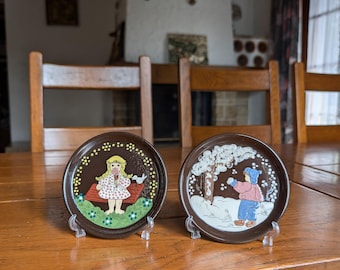 May include: Two brown decorative plates with colourful illustrations. One plate features a girl on a bench, the other a child in the snow. Both plates are displayed on clear stands, set on a wooden table.