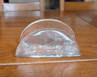 Op de afbeelding: Transparante glazen servethouder met een getextureerd oppervlak. De houder heeft een halfrond ontwerp met een platte basis. De tekst "Glasi Hergiswil" is zichtbaar op de voorkant. Een decoratief item voor de tafel.