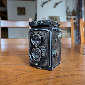 May include: A vintage Rolleiflex camera with a black body and dual lenses. The camera has the brand name "Rolleiflex" on top and "COMPUR" on the lens. The camera is sitting on a wooden table.