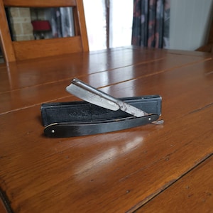 May include: An open straight razor with a dark handle and a silver blade rests on top of a black case. The razor is positioned on a wooden table, showcasing its classic design and potential use for grooming.