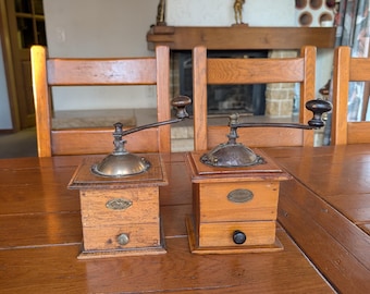 May include: Two antique wooden coffee grinders with brass tops and crank handles sit on a wooden table. The grinders have small drawers and are displayed in front of a fireplace. The overall aesthetic is vintage and rustic.