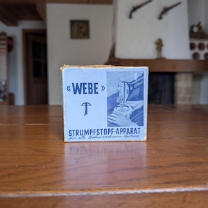 Puede incluir: Una pequeña caja azul claro con la palabra «WEBE» en letras grandes y estilizadas. La caja presenta un gráfico de una máquina de coser y el texto «STRUMPFESTOPF-APPARAT». La caja está sobre una superficie de madera.