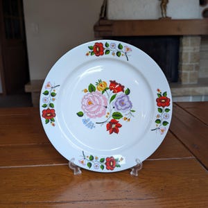 Op de afbeelding: Witte keramische plaat met een bloemmotief. De plaat heeft een centraal boeket van roze, gele en paarse bloemen, omringd door kleinere bloemclusters in rood en wit. De plaat wordt gepresenteerd op een transparante standaard.