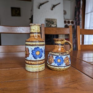 May include: Two ceramic vases with floral designs. The taller vase is cylindrical, while the smaller one has a handle. Both feature blue and brown flowers on a white background, with a brown and tan color scheme.