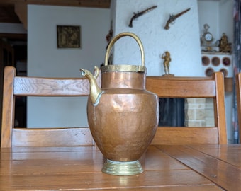 May include: A copper pitcher with a brass handle and base sits on a wooden table. The pitcher has a spout and a rounded body. The background includes a fireplace, chairs, and decorative items.