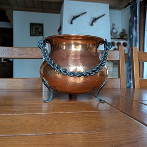 May include: A hammered copper cauldron with a dark metal chain handle and three curved legs. The pot has a shiny, reflective surface and sits on a wooden table. The background includes a fireplace and wooden ceiling.