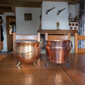 May include: Two copper pots sit on a wooden table. The pot on the left has a brass handle and three brass feet. The pot on the right has ornate handles and legs. The background includes a fireplace and decorative items.