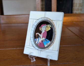 May include: A rectangular, off-white picture frame with an oval-shaped image of a woman in a pink dress and hat. The frame has a decorative border and is propped up on a clear plastic stand. The image is set against a dark background.