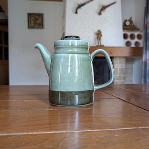 Può includere: Una teiera in ceramica con smalto verde e marrone. La teiera ha un beccuccio, un manico e un coperchio. La teiera è su un tavolo di legno.