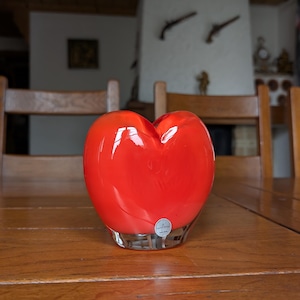 Peut inclure: Un vase en verre rouge vif en forme de cœur. Le vase a une finition brillante et repose sur une table en bois. La base transparente du vase est visible. L'arrière-plan comprend des chaises en bois et un mur blanc.