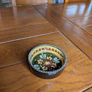 Pode incluir: Um pequeno prato redondo de cerâmica pintado à mão com exterior castanho escuro. O interior apresenta uma base verde com um padrão floral em vermelho, amarelo e branco. A borda é amarela com pontos vermelhos e uma borda branca.