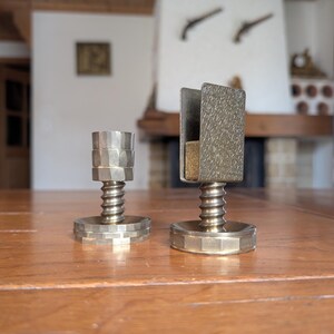 Könnte beinhalten: Zwei Messing-Kerzenhalter mit einzigartigem Design. Einer hat die Form eines traditionellen Kerzenständers, der andere hat eine rechteckige Oberseite. Beide haben eine gewindeförmige, federartige Mittelsäule und einen sechseckigen Sockel. Die Kerzenhalter stehen auf einem Holztisch.