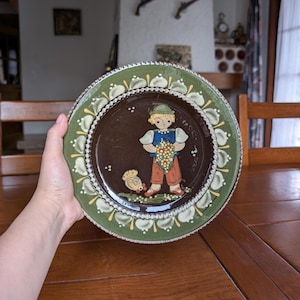 Op de afbeelding: Een decoratieve keramische plaat met een centrale illustratie van een jonge persoon die druiven vasthoudt. De plaat heeft een donkerbruin midden, een groene rand met witte en gele bloemaccenten en een geschulpte rand. De plaat wordt door een hand vastgehouden.