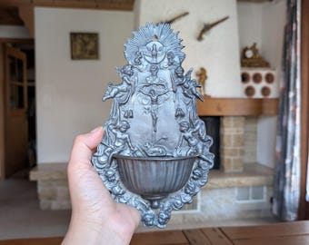 May include: A silver-colored wall-mounted holy water font with a detailed religious design. The font features a central depiction of Jesus on the cross, surrounded by angels and ornate embellishments. The bowl is at the bottom.