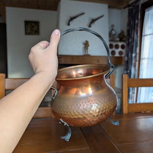 Puede incluir: Un caldero de cobre con textura martillada, con un asa curvada y tres patas decorativas. La olla tiene una superficie brillante y reflectante y la sostiene una persona. El fondo incluye una mesa de madera y una chimenea.