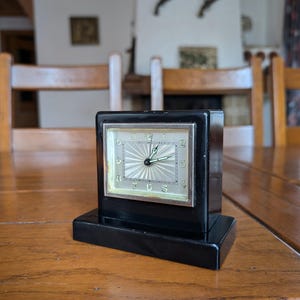 May include: A black vintage clock with a rectangular face and base. The clock face is silver with black numerals and hands. The clock is set on a wooden table.