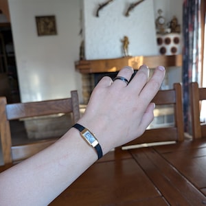 Op de afbeelding: Een vintage rechthoekig horloge met een goudkleurige kast en een zwarte gevlochten band wordt om een pols gedragen. De wijzerplaat van het horloge is licht van kleur. Een zwarte ring zit om een vinger. De achtergrond omvat een houten tafel en een open haard.