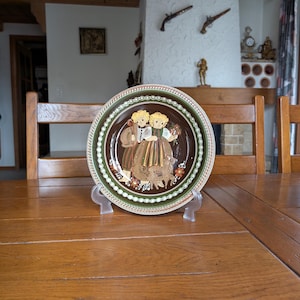 Op de afbeelding: Een decoratieve bord met een traditioneel ontwerp. De bord heeft een bruin midden met een gedetailleerde illustratie van een paar in traditionele klederdracht, omgeven door een groen en wit patroon. Het wordt gepresenteerd op een transparante standaard.