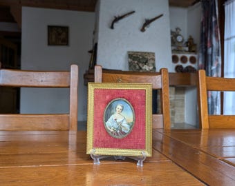 May include: A framed miniature portrait of a woman in a gold frame with a red fabric backing. The portrait is oval-shaped and features a woman holding flowers. The frame is displayed on a wooden table.