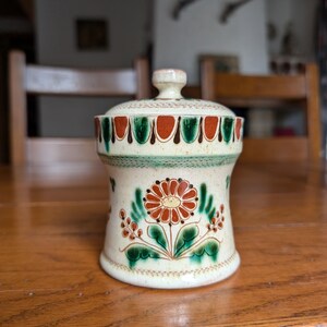 Pode incluir: Um pote de cerâmica creme com tampa, apresentando um design floral em vermelho, verde e castanho. O pote tem uma forma arredondada, uma borda decorativa e um pequeno botão na tampa. Está sobre uma mesa de madeira.