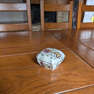 May include: A small, heart-shaped trinket box with a white base and floral design in pink, yellow, and red. The box has a gold trim and is sitting on a wooden table.