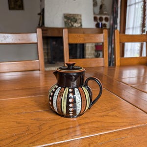 Pode incluir: Jarro de cerâmica decorativo pequeno com tampa e alça marrom escuro, e corpo estampado. O corpo apresenta listras verticais em creme, laranja e preto, com pontos brancos e detalhes de linhas onduladas. O jarro está sobre uma mesa de madeira.