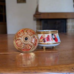 May include: An open trinket box with a lid. The lid is red and gold with an ornate design. The box is white with a gold rim and gold feet. The box has a painted design of toys and characters.