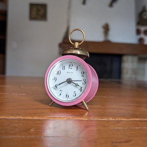May include: A retro pink alarm clock with a gold-coloured bell and legs. The clock face is white with black numerals and hands. The clock is set on a wooden surface.