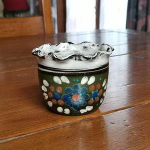 Puede incluir: Una pequeña maceta de cerámica pintada a mano con borde ondulado. La maceta presenta una base verde oscuro adornada con diseños florales azules, rojos y blancos. El borde es blanco con detalles negros, creando una estética rústica y artesanal.