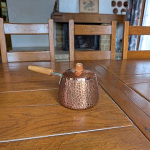 Puede incluir: Una pequeña olla de cobre martillado con asa y tapa de madera. La olla tiene un cuerpo redondeado y una superficie texturizada. El asa está sujeta con alambre. La olla está sobre una mesa de madera.