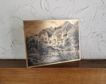 May include: A framed print with a gold border, depicting a black and white landscape of a European village in a valley. The print shows mountains, buildings, and the word "Burglen" in the upper left corner.