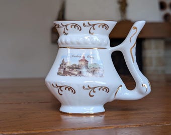 Puede incluir: Taza de cerámica blanca con un asa única y detalles dorados. La taza presenta una imagen escénica y el texto "Karlsbad Vary". Adornos dorados decoran el borde y la base. La taza se muestra sobre una superficie de madera.