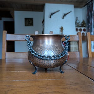 Puede incluir: Un caldero de cobre con acabado martillado, cadena decorativa y tres patas pequeñas. El caldero tiene dos asas y una tapa. El texto "25 SMU im" es visible. Está sobre una mesa de madera.