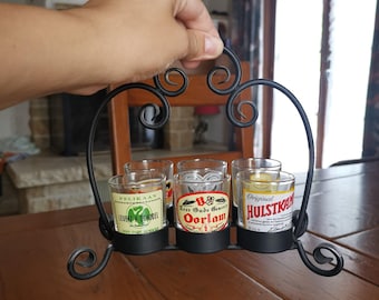 May include: A black metal shot glass holder with a decorative, scrolled design, holding five shot glasses. The shot glasses have colorful labels with text. The holder is being held by a person.