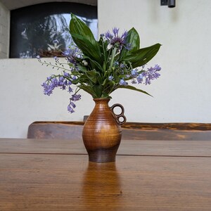 May include: A small, brown ceramic vase with a handle holds a bouquet of purple and green flowers. The vase sits on a wooden table. The flowers include bluebells and other greenery. The background is a white wall with a window.