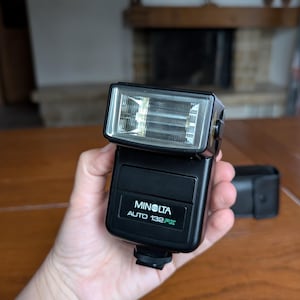 May include: A black Minolta Auto 132PX flash unit. The flash has a rectangular head with a clear front and a black body. The text "MINOLTA AUTO 132PX" is printed on the front. A black leather case is visible in the background.