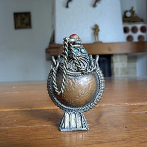 May include: An antique, ornate metal perfume bottle with a detailed, engraved copper body. The bottle features a silver-colored filigree top with a red stone and turquoise accents. A decorative chain hangs from the top. The bottle sits on a silver-colored base.