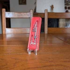 May include: A red plastic case containing a silver and black Kern Swiss compass set. The compass is displayed on a clear plastic stand on a wooden table. The case has the Kern Swiss logo.