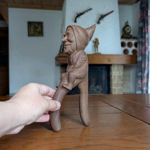 Könnte beinhalten: Ein Nussknacker aus Holz in Form eines lächelnden Gnom mit spitzer Mütze und Kapuzenmantel. Der Gnom sitzt auf den Griffen des Nussknackers, die von einer Hand gehalten werden. Der Nussknacker ist aus dunkelbraunem Holz.