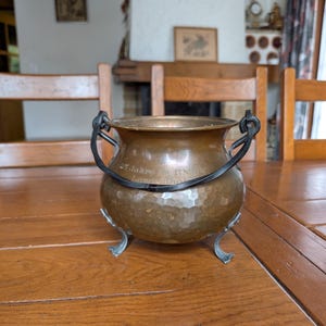 May include: A copper cauldron with a hammered finish, featuring black iron handles and feet. The cauldron has a dark patina and a rounded shape. The text "25 Jahre SN" is engraved on the rim. It sits on a wooden table.
