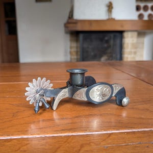 May include: A vintage metal spur with a floral design. The spur features a silver-colored, star-shaped rowel and a black strap with silver accents. The spur is resting on a wooden surface.